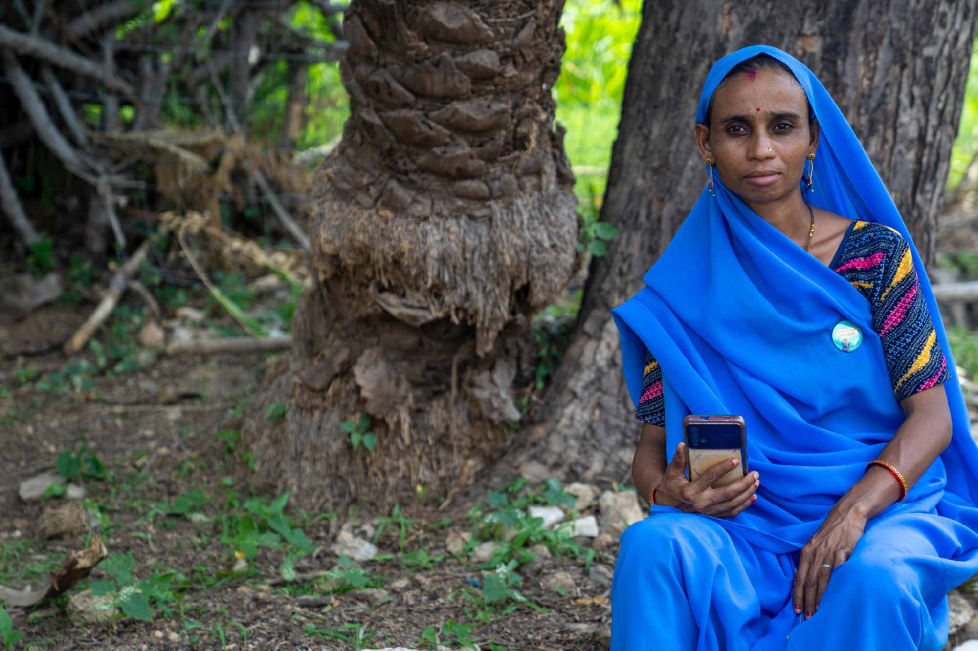 Lady in blue sari, holding a smartphone, looking at the camera. Seated outside in front of a couple of trees.