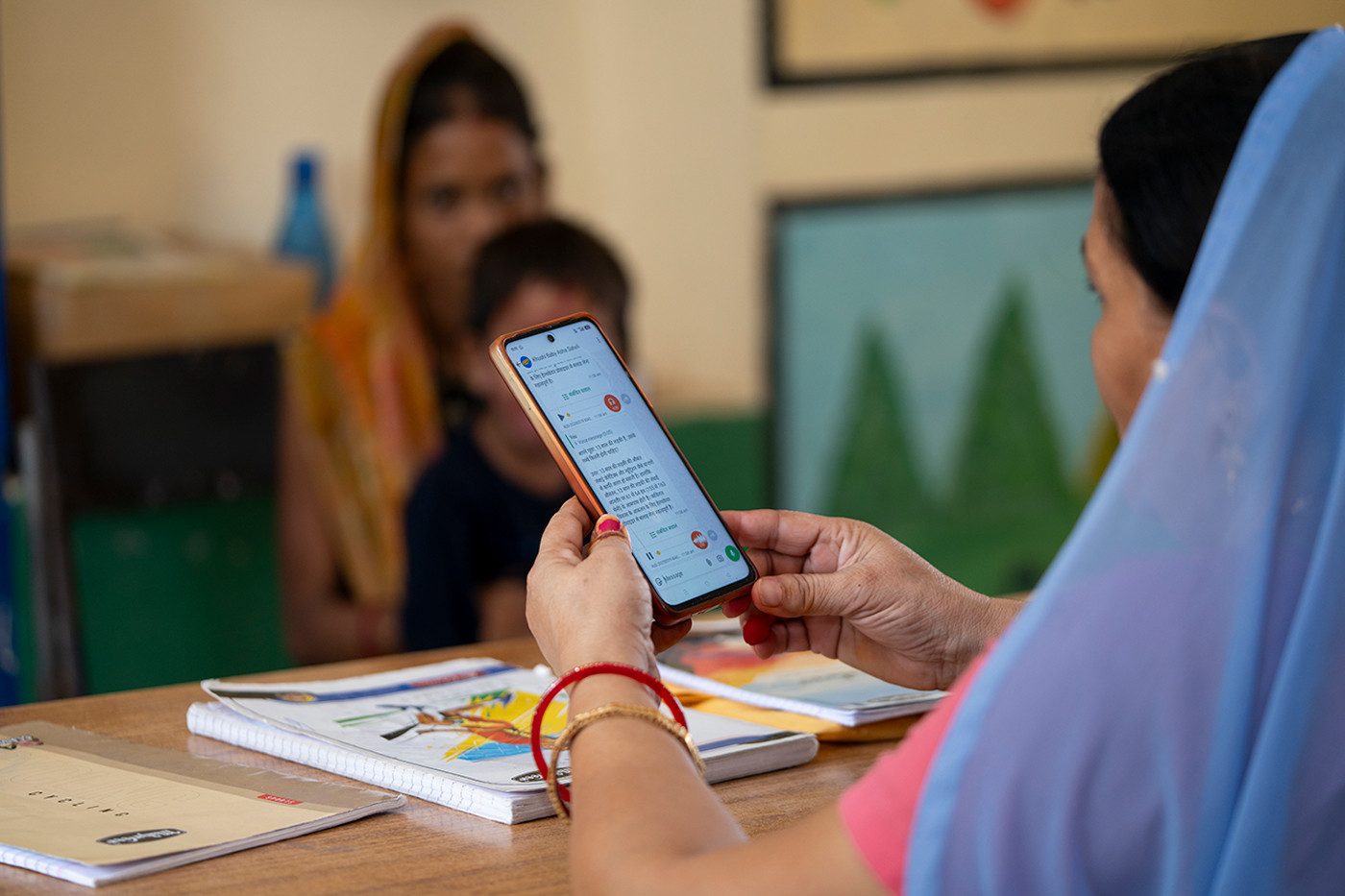 The back of the head of a person wearing a sari holding a smartphone with Hindi writing, talking to a mother and child who are blurred in the background.
