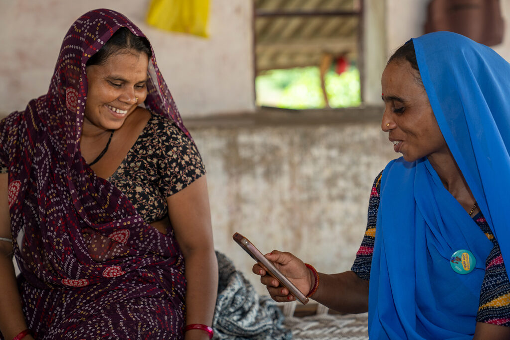 One lady in a maroon sari talking to a lady in blue sari, holding a smartphone, sitting in a white room.