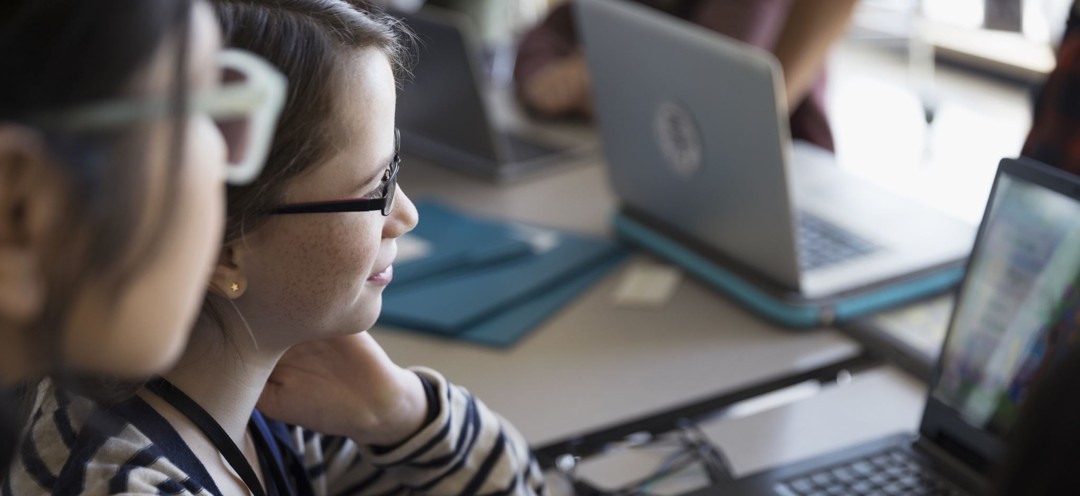 Two female middle-school students seated at a table, focused on a laptop screen. The girl in the foreground wears glasses and a striped shirt, typing with one hand while the other rests on their neck. A second girl observes from beside her. The table holds additional laptops and folders, suggesting a collaborative or educational setting. The scene conveys active engagement with technology in a learning or workshop environment.