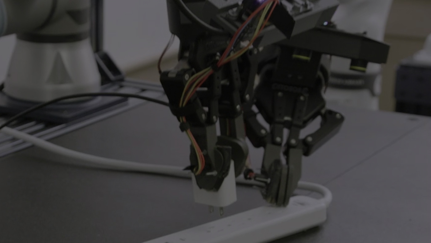 A dual-arm robot with two-finger grippers is unplugging a power adapter from a white power strip on a workbench.