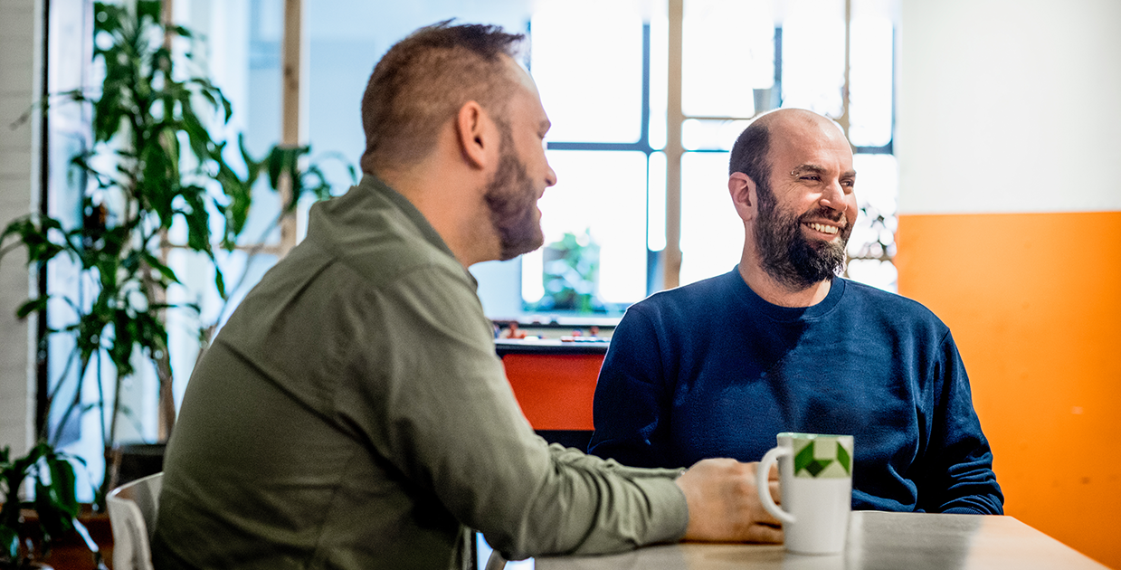 Two workers sit smiling at a table, over a cup of coffee.