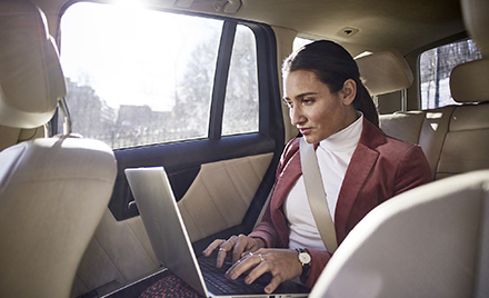 A woman in a car types on her laptop.