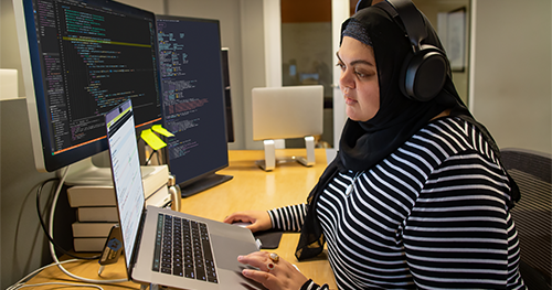An image of a female developer coding at her desk, wearing hijab and headphones.