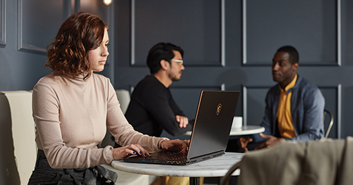 A remote female tech worker on her laptop while two co-workers have a discussion in the backgroud.