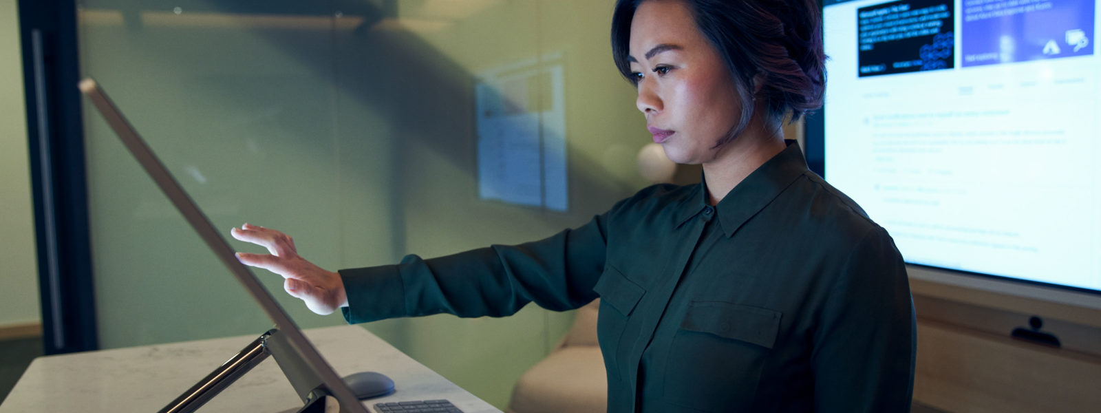 Side profile of a woman wearing a dark shirt in a dim office scrolling or working on a Microsoft Surface Studio.