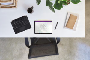 Bird‘s eye view of Surface Pro X projecting OneNote with a detached black keyboard, pen, books, plant, mug, and wooden shelf.