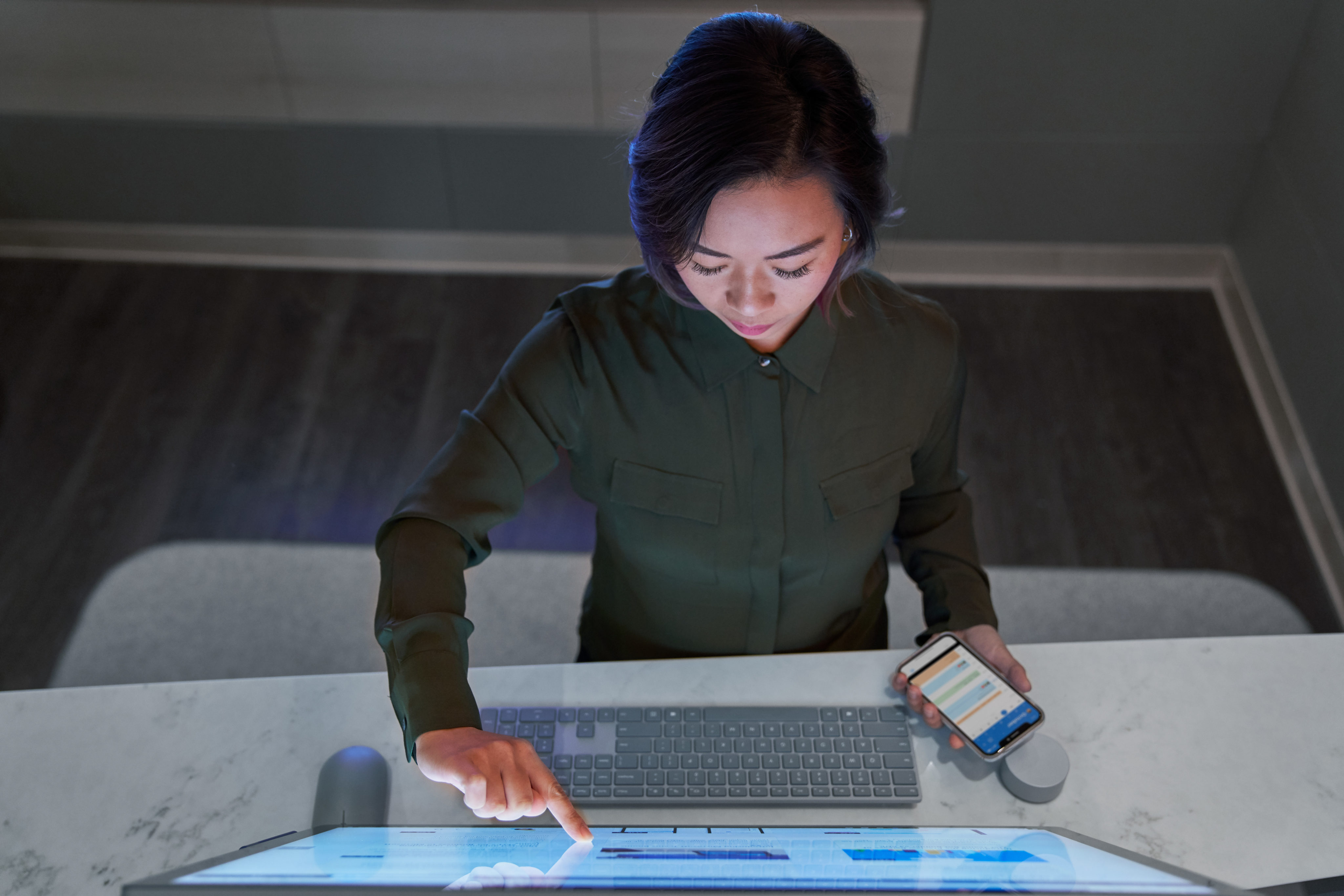 Top down view of a woman wearing a dark shirt in a dim office scrolling or working on a Microsoft Surface Studio and holding a phone.