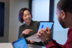Smiling woman in a meeting room holding up a Surface laptop presenting statistics. Blurred man in foreground.