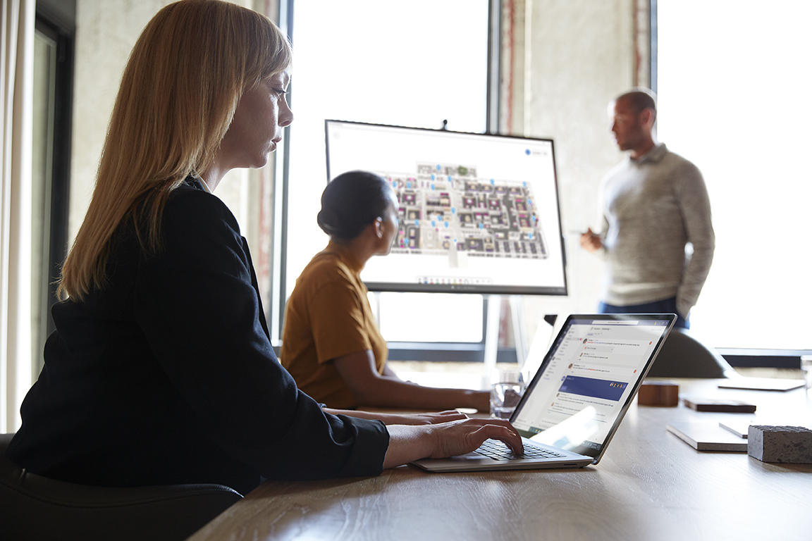 Three adults are collaborating in a conference room. One male is standing next to a Microsoft Surface Hub 50” with Microsoft Whiteboard screen shown.