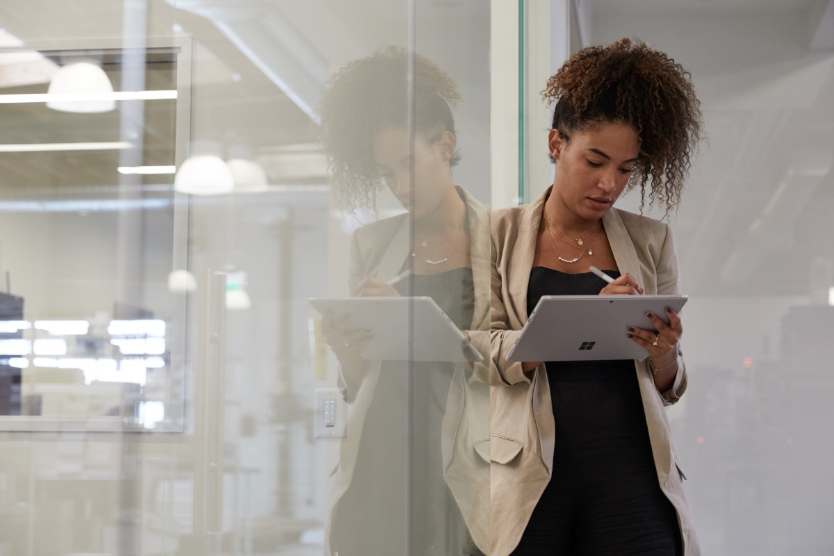 Adult female in industrial office setting leaning against a glass wall while holding a platinum Microsoft Surface Pro 7 in tablet mode preparing to write with Microsoft Surface Pen.