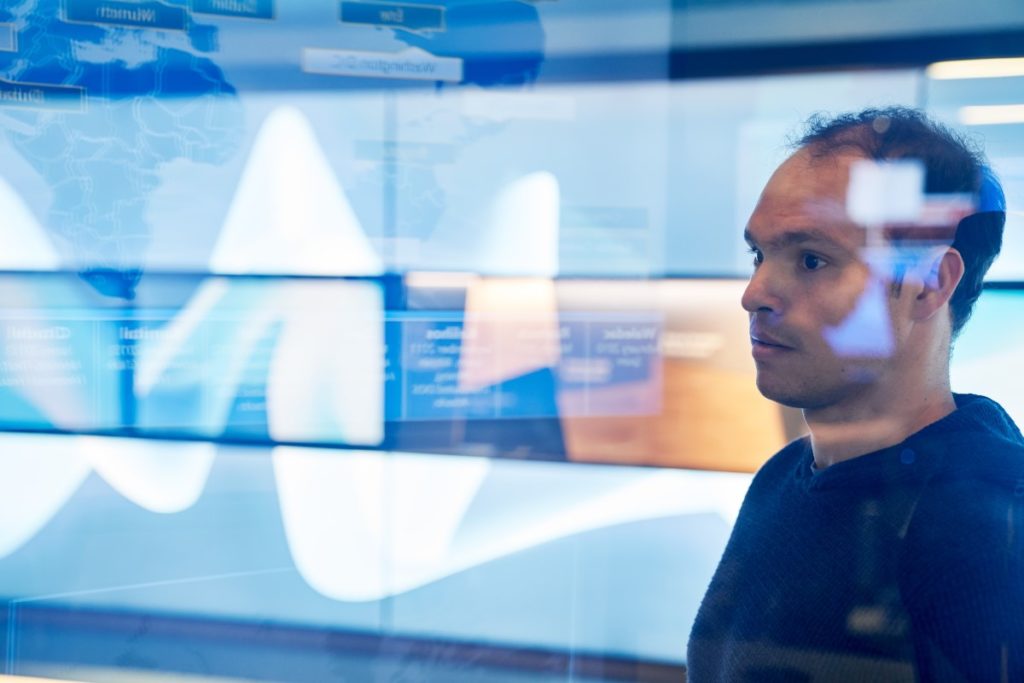 Man in hooded sweater/sweatshirt inside a secure room who is looking at data and a geographic area displayed on a large monitor which is behind glass walls with reflections.
