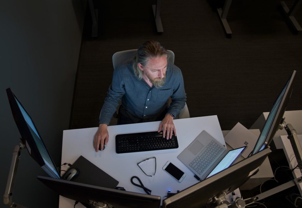 Top-down view of a bearded man in a gray/blue shirt seated at a desk working on a Surface laptop connected to three large monitors. Desktop has phone, glasses, mouse and other laptops on it.