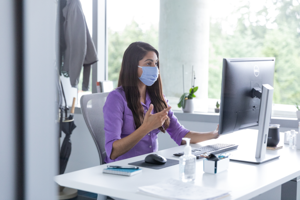 A female employee wearing a mask and talking on a video call with hand sanitizer on her desk, socially distancing from others.