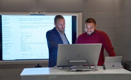 Forward facing view of two men working on a Microsoft Surface Studio with a larger blurred screen/display behind them.