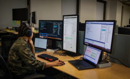 Black female developer wearing headphones; coding at her PC workspace in an enterprise office, using Visual Studio on a multi-monitor set up.