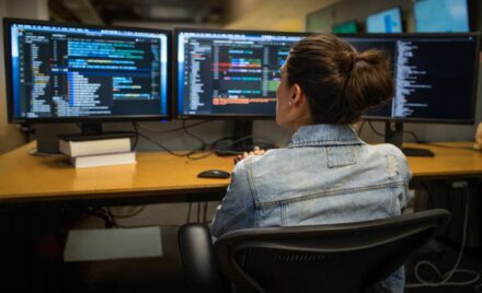 Female developer coding her workspace in an enterprise office, using Visual Studio on a multi-monitor set up.
