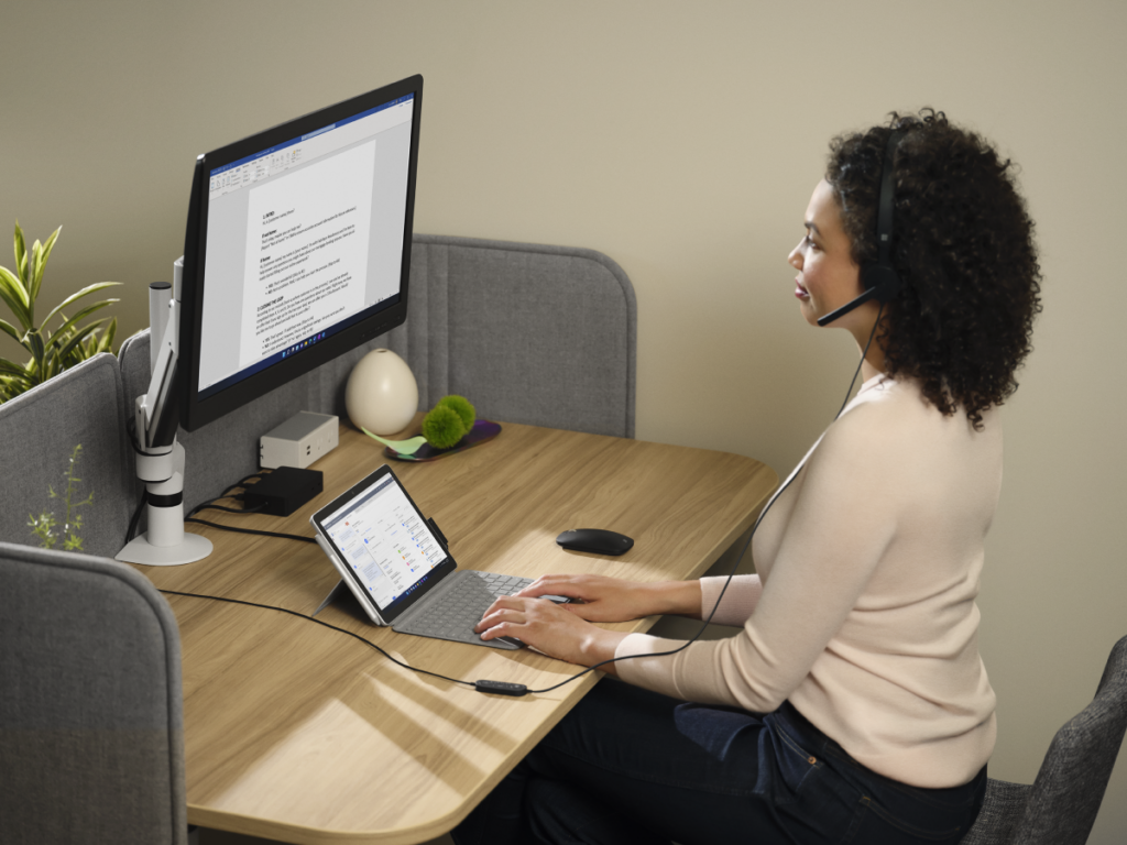 Adult female wearing Microsoft Modern USB-C Headset sitting at a desk while working on a Surface Go 3 with Surface Go Typecover in laptop mode with Microsoft Edge displayed. Microsoft Word is being presented on the large monitor.