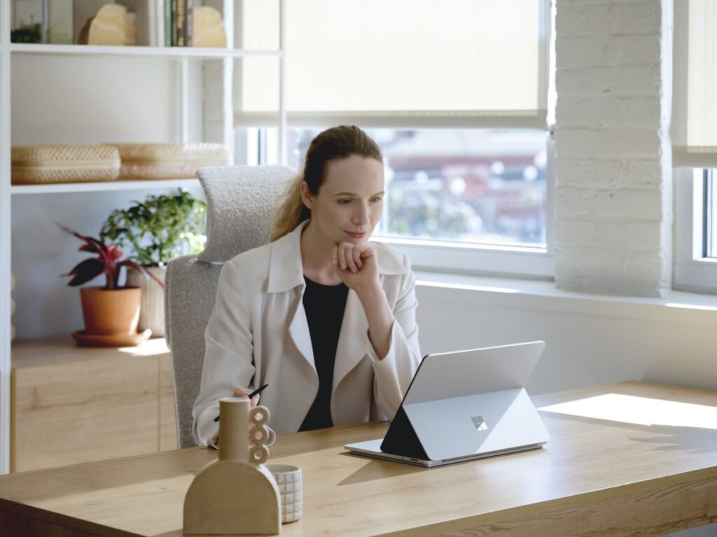 Adult female sitting at a desk, holding a Microsoft Slim Pen 2 while looking onto a platinum Surface Laptop Studio in stage mode.