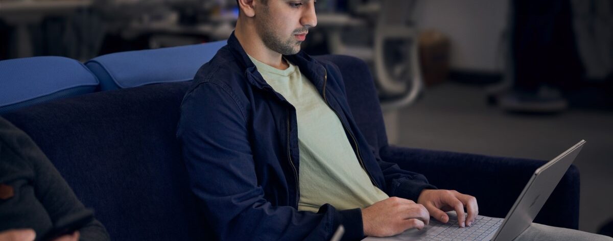 Lightly bearded man working on a laptop while sitting next to another person on a dark blue couch.