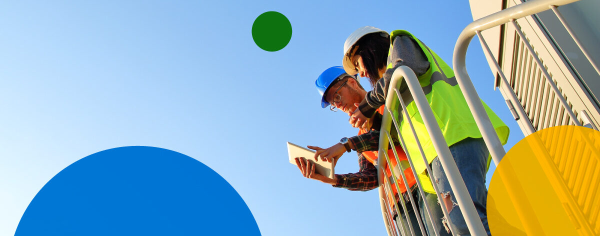 Two adults stand next to railing wearing safety vests with blue sky in background.
