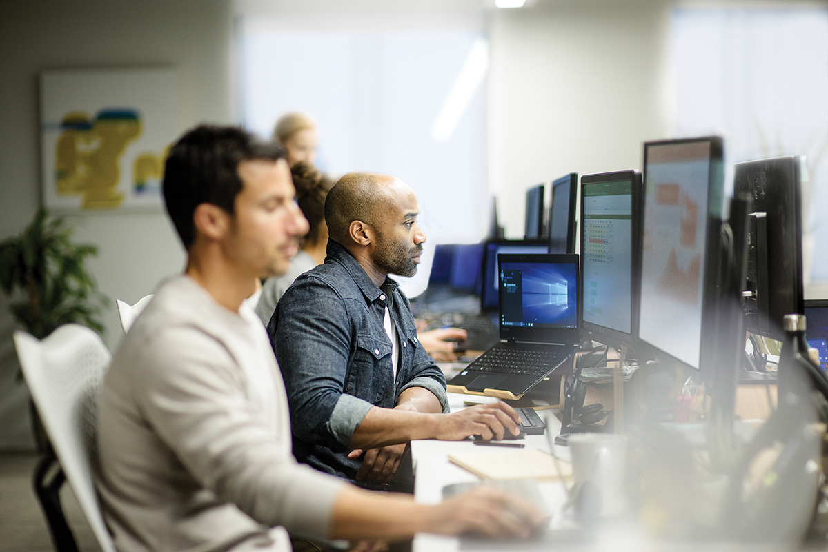 Two male coworkers sitting next to each other along row of office desks, using their own desktop computers (screens show Excel and PowerPoint). An open HP laptop sits on desk to their left, showing the Windows 10 home screen.