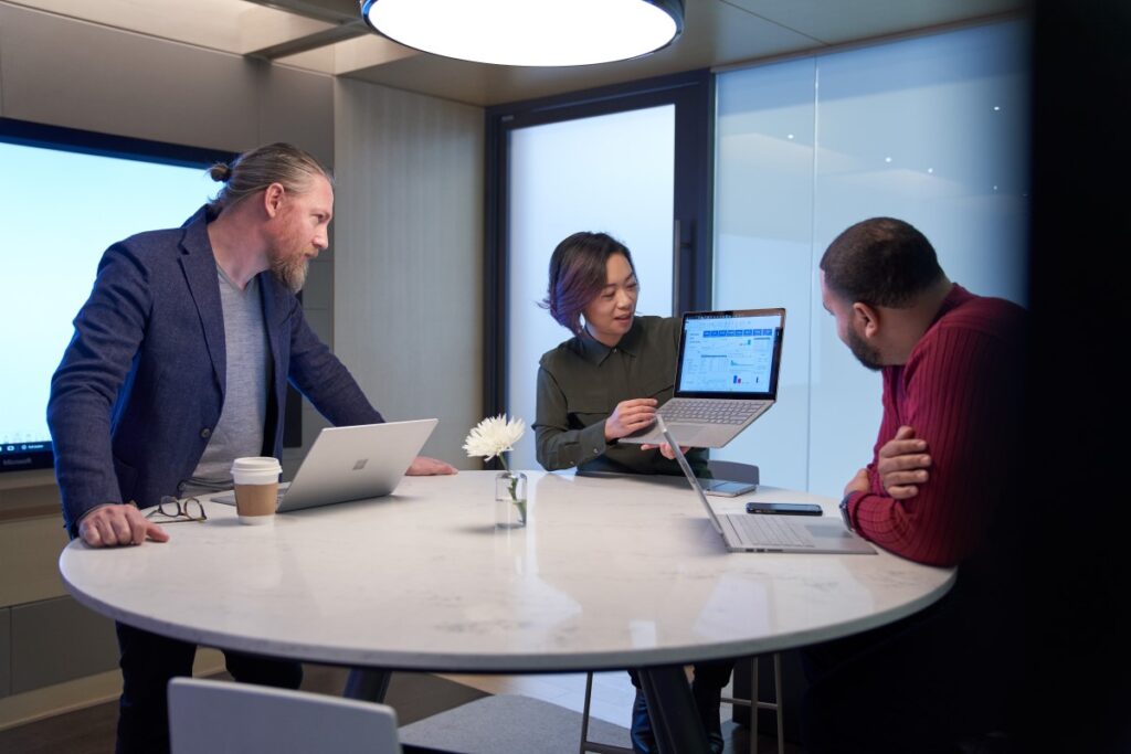 Woman and two men working in a meeting room all with Surface laptops. The woman is holding up her laptop and presenting her screen to the two men.