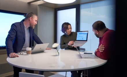Woman and two men working in a meeting room all with Surface laptops. The woman is holding up her laptop and presenting her screen to the two men.