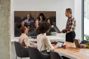 A male speaker presenting in a Microsoft Teams Room meeting while viewing remote participants in Together Mode on the front of room screen​.