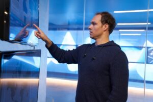 Man in a hooded sweatshirt inside a secure room, pointing at a geographic area displayed on a large monitor.