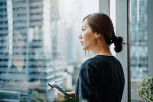 Young businesswoman holding a phone, looking out her office window overlooking the city.