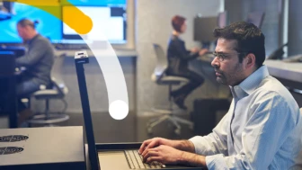 Man in a collared shirt working on a server station inside a secure room. Coworkers and large monitors are in the background.