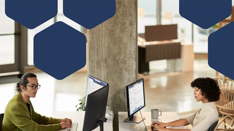 Photo of two employees collaborating during a Microsoft Teams meeting while working in an open office setting on dual monitors.