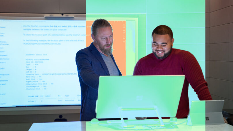 Two men look at a computer screen together.