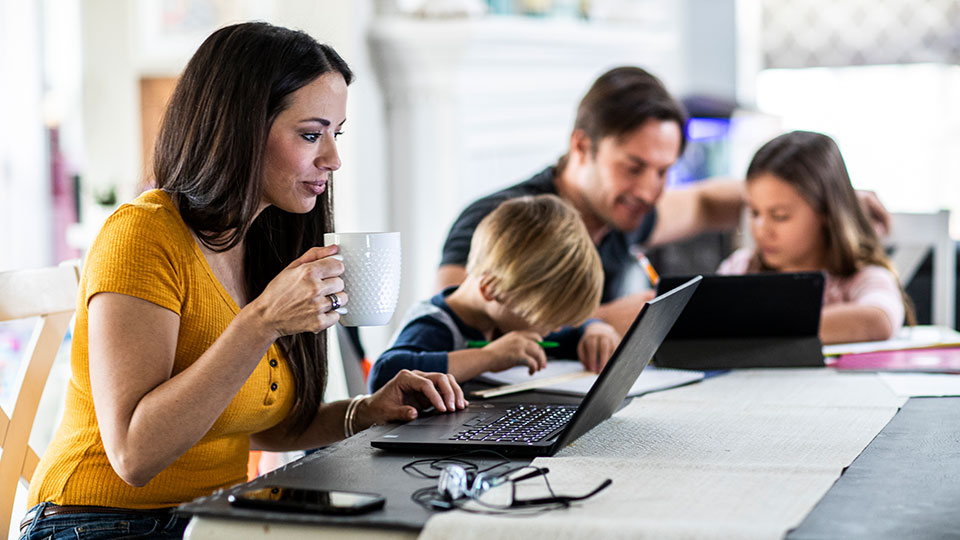 A woman works at her laptop at a kitchen table with family