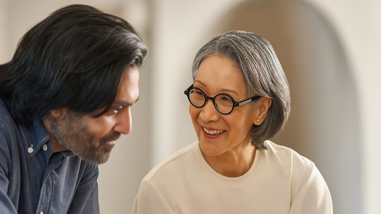 Smiling woman with glasses talking to a man with long hair indoors.
