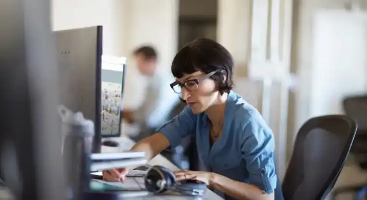 Office employee in a blue shirt working at a desktop computer in a modern open plan office, writing notes while viewing content on a large monitor.
