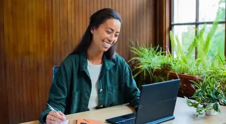 Remote worker in a green jacket sitting at a desk with a laptop, holding a pen and looking toward the screen in a bright workspace with large windows and potted plants in the background.