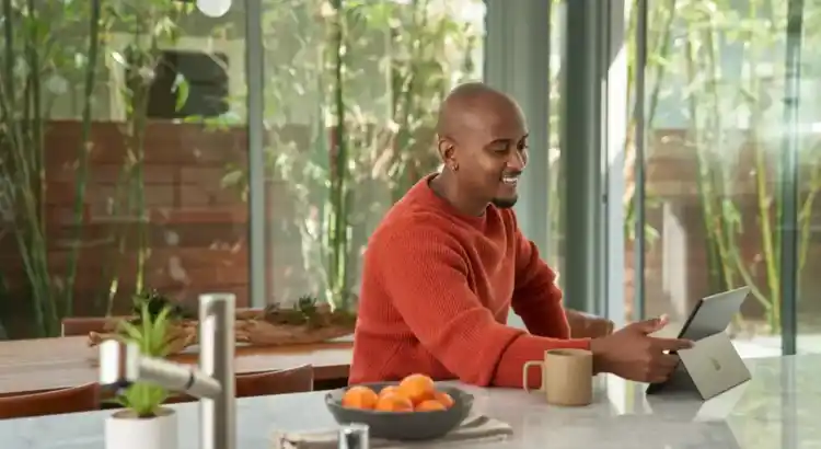 Remote worker in an orange sweater sitting at a kitchen island, using a tablet with a mug of coffee and a bowl of oranges nearby, surrounded by large windows overlooking a garden.