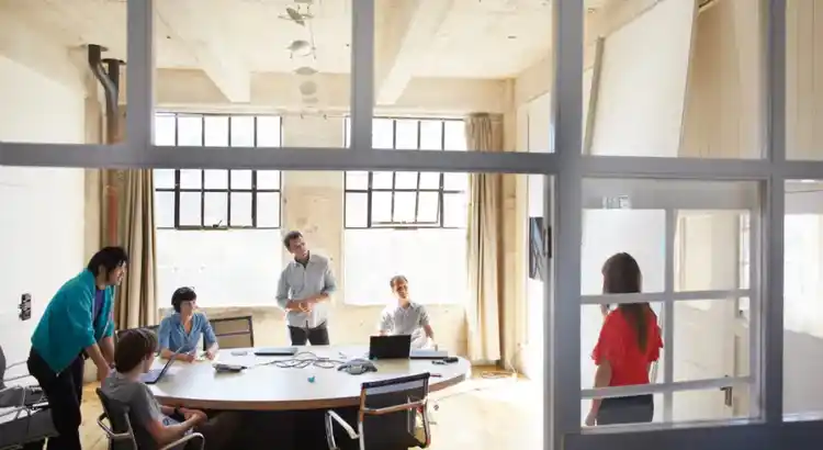 Team meeting in a bright loft style conference room, with several colleagues gathered around an oval table discussing work while another person in a red shirt enters through the glass door.