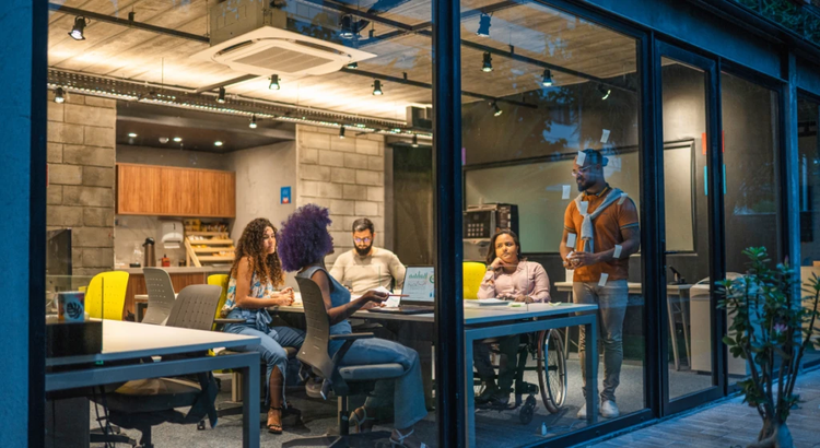 Evening view through glass walls of a modern office where a diverse group of people work and collaborate around tables, some seated and one standing and speaking.