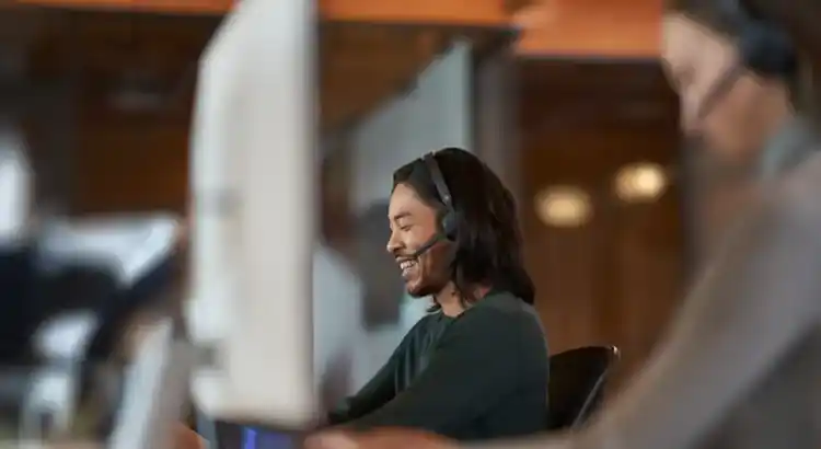 Smiling person wearing a headset sits at a desk in an office, looking at a monitor while speaking.