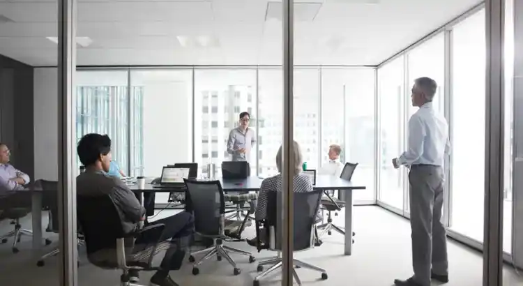 Several people sit around a conference table in a bright glass-walled meeting room, with laptops open in front of them.