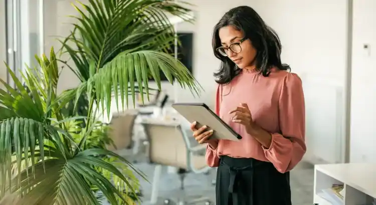 Person in a pink blouse and glasses stands in a bright office near large green plants, focused on using a tablet.