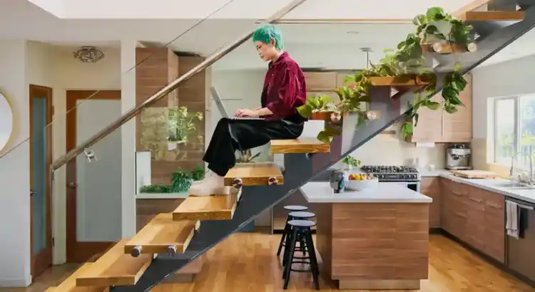 Person with bright teal hair sits cross-legged on a floating wooden staircase, working on a laptop in a modern open-plan kitchen.