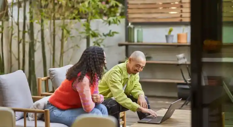 Two people sit together on outdoor lounge chairs in a garden-like patio, looking at a laptop on a low wooden table.