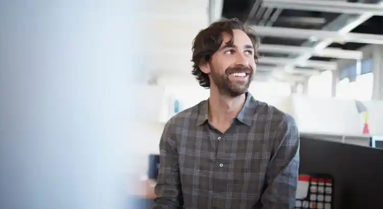 Smiling person with wavy hair and a beard wearing a plaid shirt, sitting in a bright open-plan office.