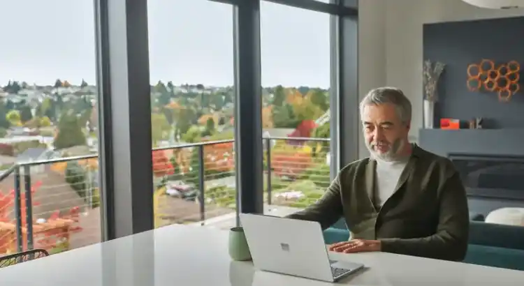 Person with gray hair and beard sitting at a white table, working on a laptop in a modern home with large windows overlooking a suburban landscape.