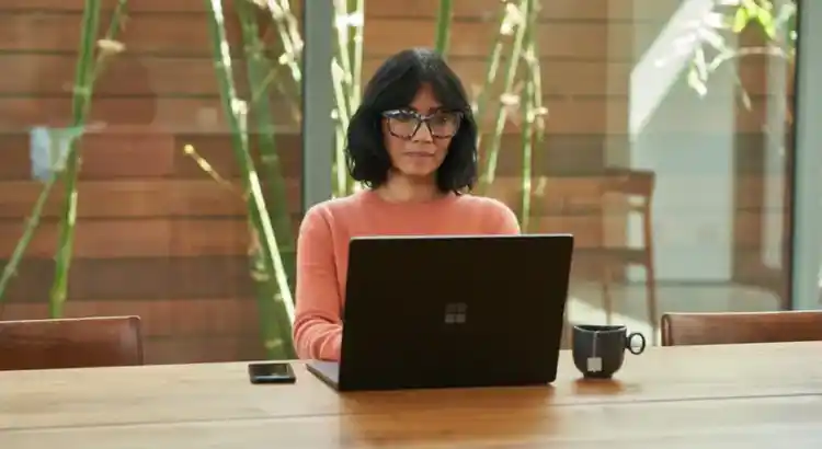 Person wearing glasses and an orange sweater working on a laptop at a wooden table with a mug and phone nearby in a bright, plant-filled space.