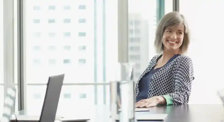Smiling person sitting at a conference table in a bright office, turning slightly in their chair with an open laptop nearby.